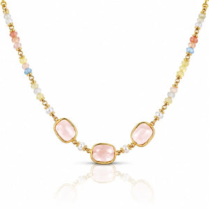 A gold-tone beaded necklace featuring three central light pink rectangular gemstones, flanked by small white pearls and multicolored beads in pastel shades of yellow, blue, and peach, set against a clean white background with a soft reflection below.
