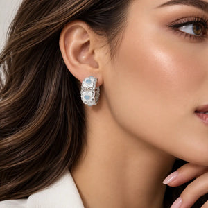 A close-up of a woman's ear wearing a silver-toned hoop earring with three oval light blue gemstones and a halo of small clear stones.