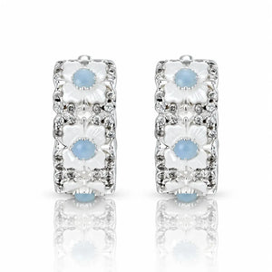 A pair of silver huggie hoop earrings decorated with three stacked white floral motifs, each featuring a light blue cabochon stone and surrounded by small clear crystals, set against a white background with a soft reflection below.