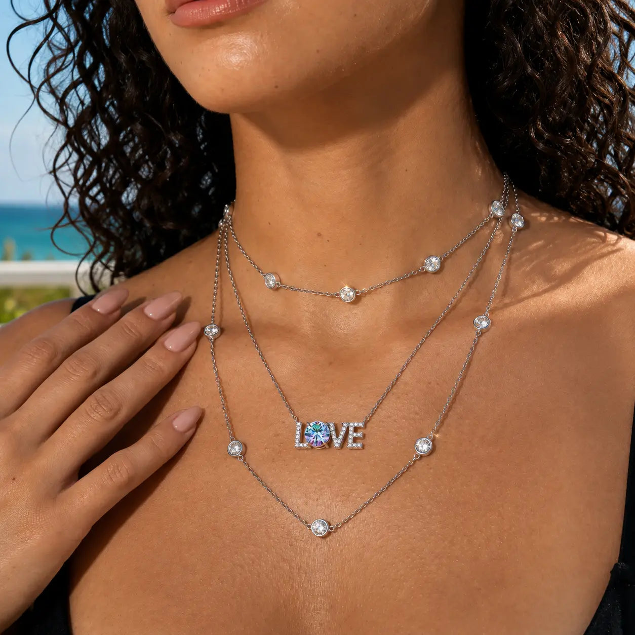 A close-up lifestyle shot of a model wearing a layered silver necklace set. The set includes a short chain with round clear stones, a longer chain with a 'LOVE' pendant featuring a blue heart in the center, and a delicate chain with a single clear stone.