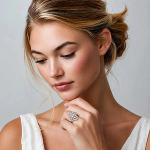 A close-up lifestyle shot of a woman with blonde hair in an updo, wearing a large, intricate silver ring with a flower-like design set with clear stones. She is wearing a white top against a neutral gray background.