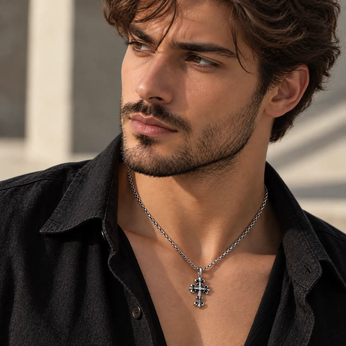 A close-up of a man wearing a silver-toned cross pendant necklace over a black shirt, photographed in warm natural light against a softly blurred outdoor background.