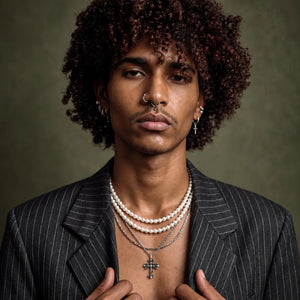 A close-up portrait of a man with curly dark hair wearing layered jewelry, including a silver cross pendant necklace, a pearl strand necklace, and a silver chain necklace, over a dark pinstripe blazer.