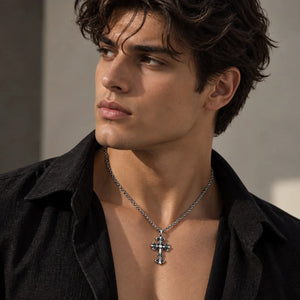 A close-up of a man wearing a silver cross pendant necklace with clear crystals, styled with a black open-collar shirt against a neutral gray background.