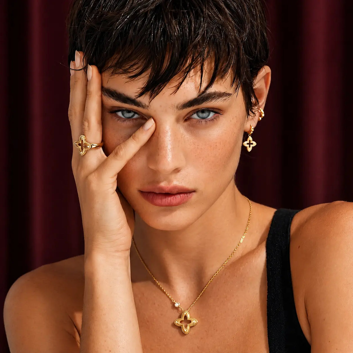 A close-up portrait of a model wearing gold jewelry, including a gold necklace with a clover-shaped pendant, a matching ring, and earrings, set against a dark red curtain background.