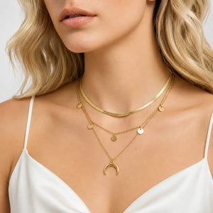 A close-up of a woman wearing a layered gold necklace set, featuring a choker, a chain with small disc charms, and a longer chain with a crescent moon pendant, styled with a white camisole top.