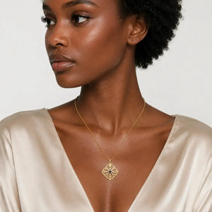 A close-up of a woman wearing a delicate gold chain necklace with a geometric diamond-shaped pendant featuring a dark blue center, styled with a satin champagne-colored top against a light gray background.
