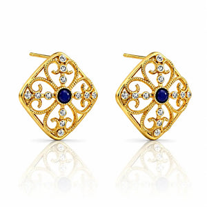 A pair of gold filigree stud earrings with a central round blue gemstone and small clear rhinestone accents, shown on a white background with a soft reflection below.