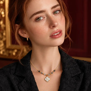A close-up portrait of a woman wearing a delicate gold necklace with a white four-leaf clover pendant and small gold hoop earrings, set against a warm, dark red and gold background.