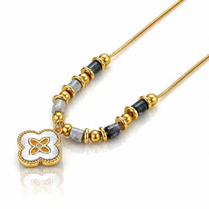 A gold-plated beaded necklace with a clover-shaped pendant. The necklace features a gold chain, gold spacer beads, gray stone beads, and a gold clover pendant with a white enamel center and gold floral details.