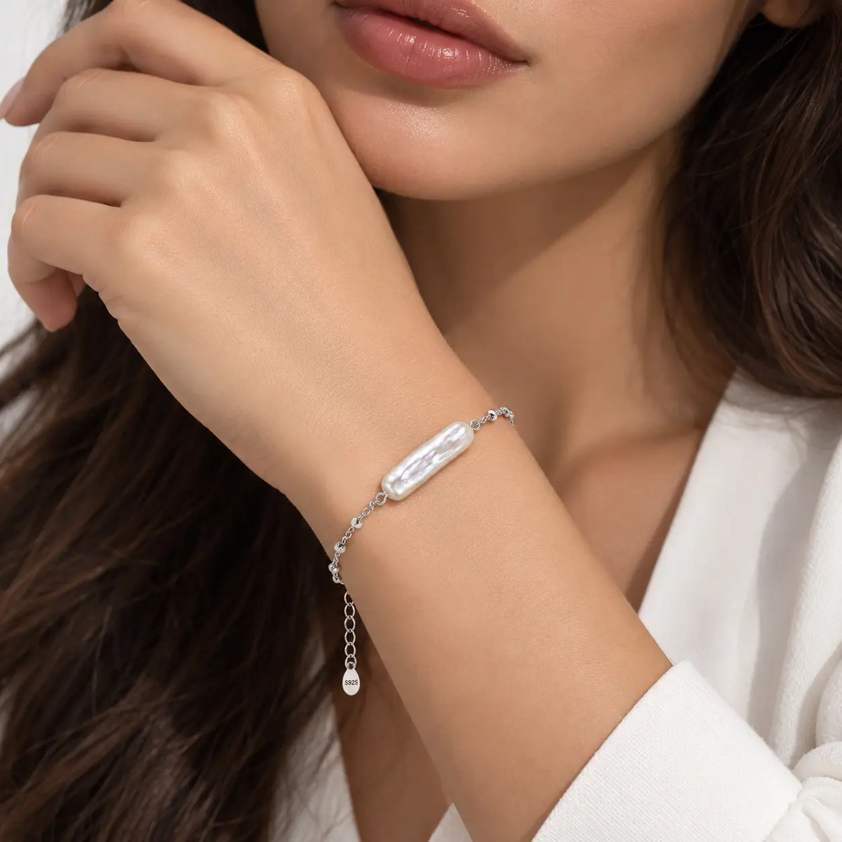 A close-up lifestyle shot of a woman wearing a delicate silver chain bracelet with a textured white bar and a small dangling pearl bead.