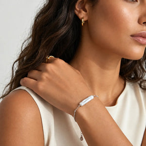 A close-up lifestyle shot of a woman wearing a delicate silver chain bracelet with a white baroque shell centerpiece, along with gold hoop earrings and a gold ring.