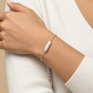 A close-up of a person's wrist wearing a delicate silver chain bracelet with a single baroque pearl pendant, paired with a cream-colored knit sweater.