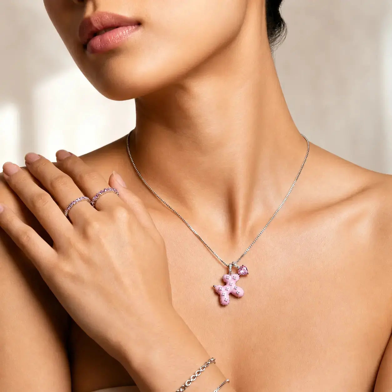 A close-up lifestyle shot of a model wearing a delicate silver chain necklace with a pink teddy bear pendant, along with matching pink and silver rings and a bracelet.
