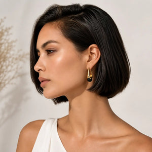 A woman with a short dark bob hairstyle is shown in profile wearing a gold and black dangle earring. She is wearing a white top against a soft, light beige background with subtle dried floral shadows.