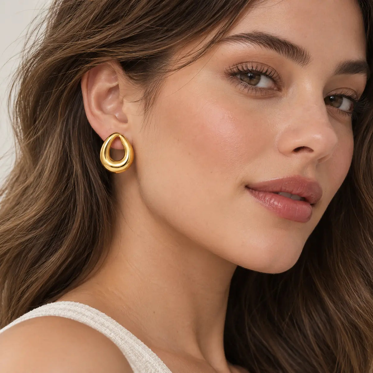 A close-up portrait of a model with short dark hair wearing gold hoop earrings with dark stone accents, against a clean light gray background.