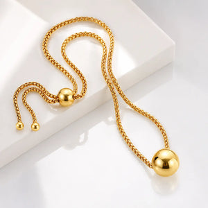 A gold-toned chain necklace with a large polished spherical pendant, displayed on a clean white geometric surface.