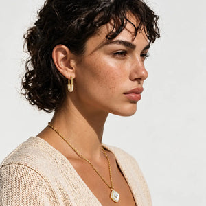 A close-up side profile of a woman with short curly dark hair wearing a delicate gold necklace with a square pendant and a matching small gold hoop earring, against a clean off-white background.