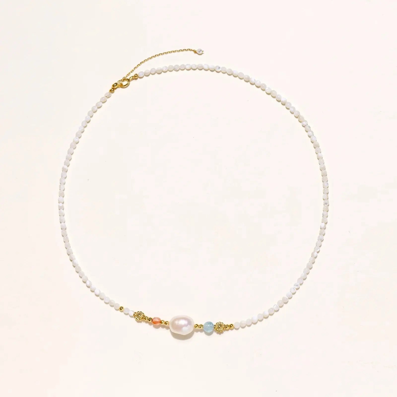 Delicate white beaded necklace with baroque pearl center, gold accents, and coral and aqua gemstones on cream background.