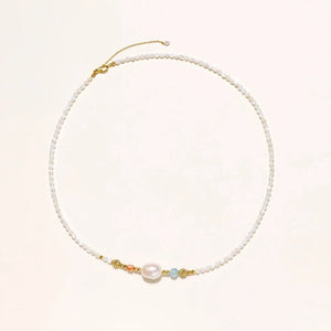 Delicate white beaded necklace with baroque pearl center, gold accents, and coral and aqua gemstones on cream background.