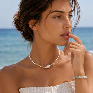 A woman on a beach wearing a delicate pearl necklace with a large central pearl and small pastel beads, along with a matching pearl bracelet.