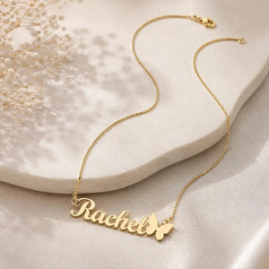 A gold nameplate necklace spelling 'Rachel' with a butterfly accent, displayed on a cream-colored stone slab with soft shadows.