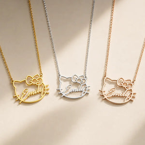 Three custom name necklaces with a Hello Kitty cat face outline pendant spelling 'Emily' are displayed on a warm beige background. The necklaces are shown in three metal finishes: gold, silver, and rose gold.