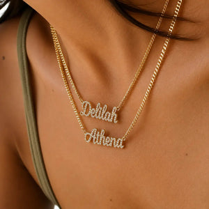 Close-up of a person wearing two layered gold necklaces with script name pendants, one reading 'Deblah' and the other 'Athena', set with small clear stones.
