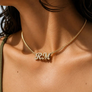 A close-up of a model's neck and chest wearing a delicate gold chain necklace with a custom 'RM' initial pendant encrusted with small clear stones.