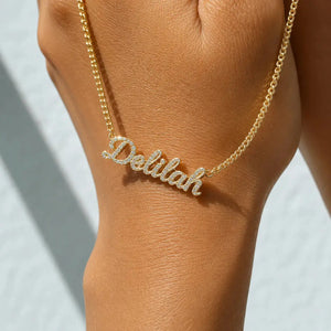 A gold nameplate necklace spelling 'Delilah' with small clear stones, worn on a person's wrist against a light gray background.