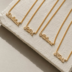 Four gold nameplates on matching chains arranged diagonally on a textured white display block. The nameplates are personalized with words like 'Daddy', 'Mom', 'Father', and 'Miss' in a script style with small clear stones.
