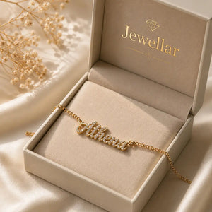 A gold nameplate necklace with small clear stones spelling 'Athena' rests inside an open beige jewelry box branded 'Jewellar'. The box is set on soft satin fabric with dried flowers in the background.