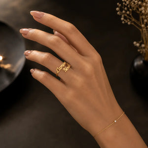 A hand with manicured nails wearing a gold ring engraved with the names 'Cassandra' and 'Nola', paired with a delicate gold chain bracelet with a small white stone, set against a dark, softly lit background.