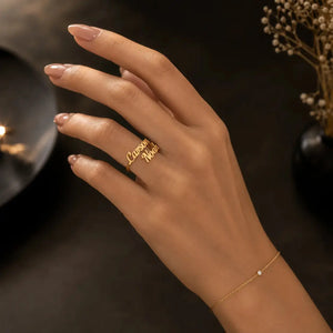 A hand with manicured nails wearing a gold ring engraved with the names 'Cassandra' and 'Nola', paired with a delicate gold chain bracelet with a small white stone, set against a dark, softly lit background.