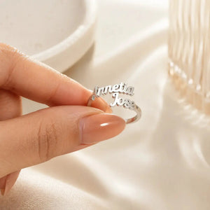 A close-up of a hand holding a silver ring engraved with the names 'Innetia Jose' on a soft, cream-colored fabric background.