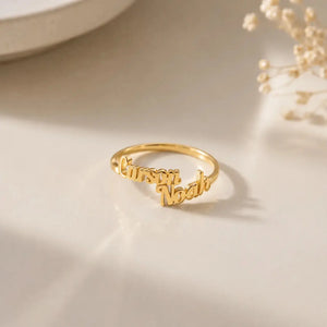 A gold name ring engraved with the names 'Cursari' and 'Noah' in cursive script, displayed on a light beige surface with soft shadows and dried flowers in the background.