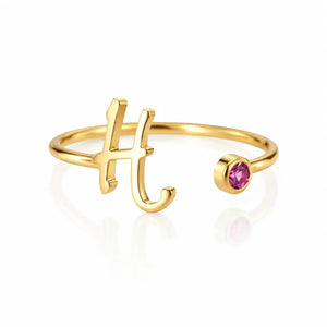 A gold initial ring featuring the letter 'H' in a polished script style, with a small round pink gemstone accent on the opposite side, set against a clean white background with a soft reflection.