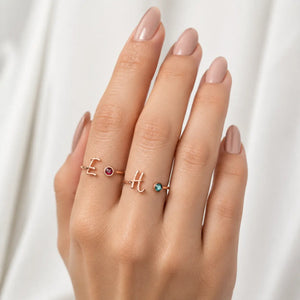 A close-up of a hand wearing three delicate rose gold rings with letter and gemstone designs, set against a soft white draped fabric background.