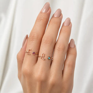 A close-up of a hand wearing three delicate rose gold rings with letter and gemstone designs, set against a soft white draped fabric background.