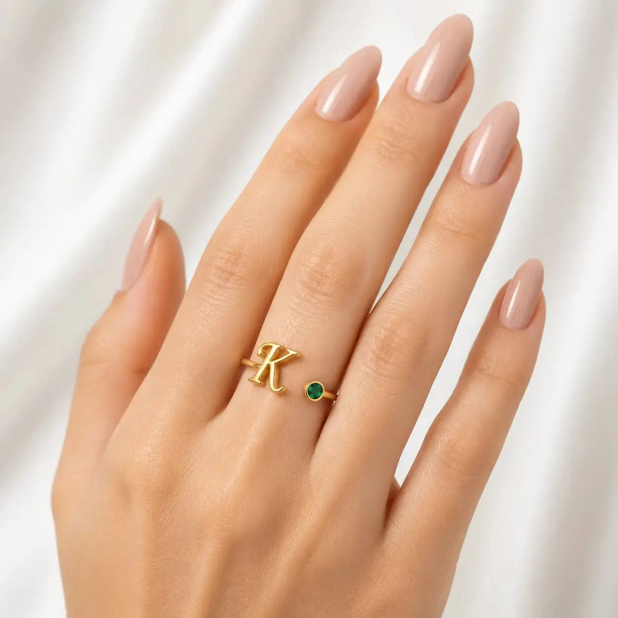 A close-up of a hand with manicured nails wearing a gold ring featuring a large letter 'K' and a small round green stone.