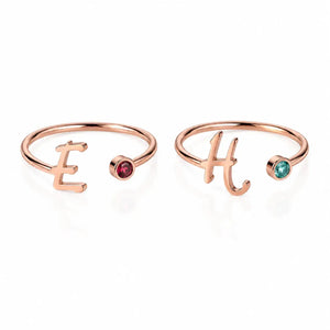 Two rose gold open bangle rings on a white background, one featuring the letter 'E' with a small red gemstone and the other featuring the letter 'H' with a small green gemstone.