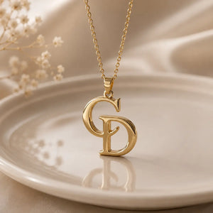 A gold initial necklace featuring interlocking letters 'C' and 'D' on a delicate chain, displayed on a warm beige ceramic plate with dried baby's breath flowers in the background.