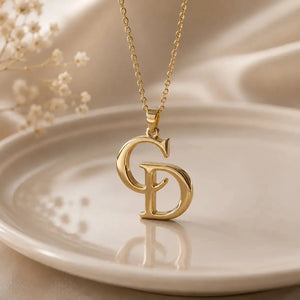 A gold initial necklace featuring interlocking letters 'C' and 'D' on a delicate chain, displayed on a warm beige ceramic plate with dried baby's breath flowers in the background.