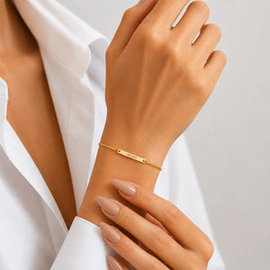 A delicate gold chain bracelet with a central bar engraved with the name 'SARAH' is shown on a model's wrist. The model is wearing a white collared shirt, and the background is a clean, neutral gray.
