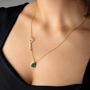 A close-up of a model wearing a delicate gold necklace with a script nameplate and a teardrop-shaped green gemstone pendant.