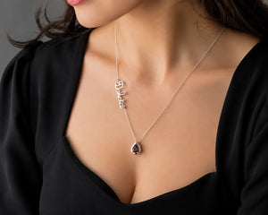 A close-up of a woman wearing a delicate silver necklace with a script name pendant and a teardrop-shaped dark red gemstone pendant, styled with a black top.