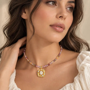 A close-up of a woman wearing a delicate beaded necklace with a gold pendant. The necklace features small, multicolored beads in shades of pink, purple, and gold, with a central gold medallion pendant containing a white stone.