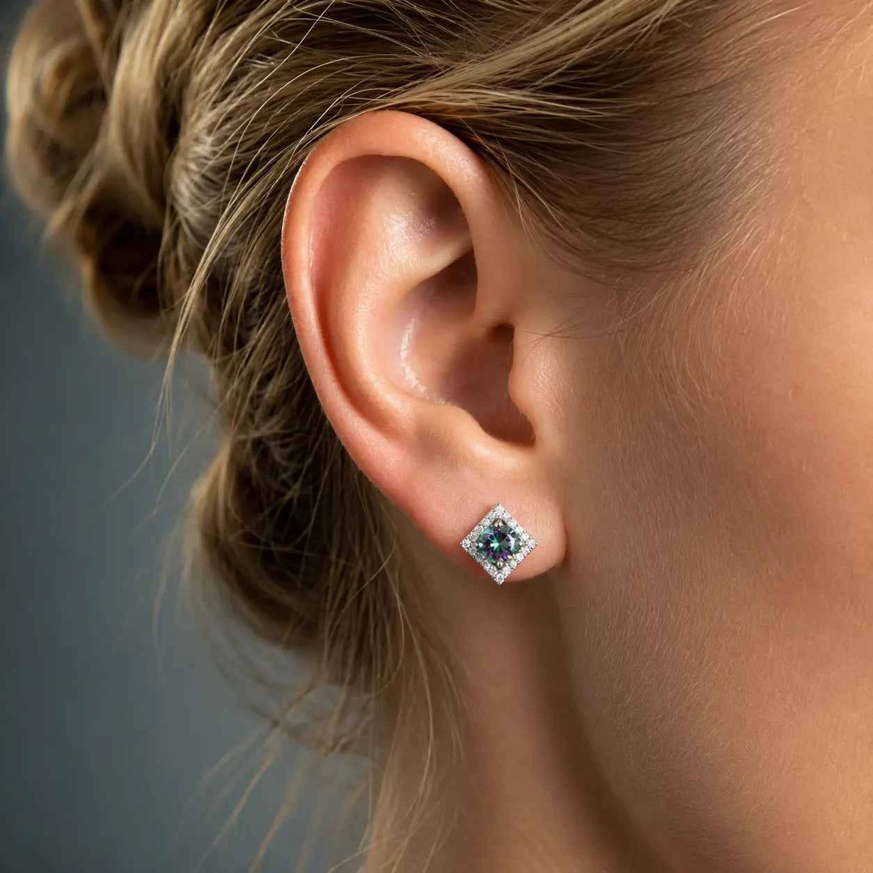 A close-up of a woman's ear wearing a small square stud earring with a dark blue center stone surrounded by clear stones in a silver-toned setting.