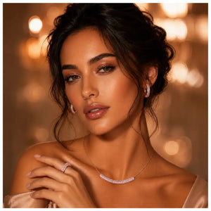 A close-up portrait of a woman wearing a delicate silver necklace with a curved pavé-style centerpiece, matching drop earrings, and a ring, set against a warm, softly blurred bokeh background of golden lights.