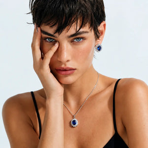 A close-up of a model wearing a silver necklace with a round blue gemstone pendant and matching blue gemstone stud earrings, styled with a black spaghetti-strap top against a light gray background.